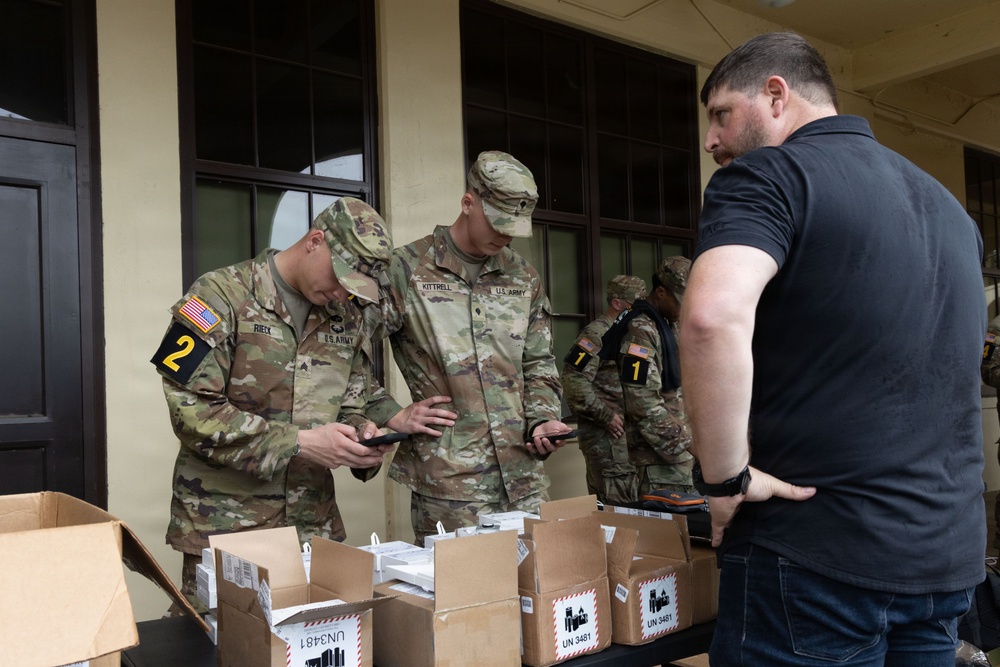 Two U.S. Army soldiers in OCP camouflage uniforms are examining items, likely smartphones or devices, from open cardboard boxes labeled 'UN 3481'. A civilian in a black shirt observes the process. The scene appears to be a distribution or inventory check of lithium battery-containing devices (UN 3481 is the UN number for lithium-ion batteries in equipment).