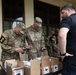 Two U.S. Army soldiers in OCP camouflage uniforms are examining items, likely smartphones or devices, from open cardboard boxes labeled 'UN 3481'. A civilian in a black shirt observes the process. The scene appears to be a distribution or inventory check of lithium battery-containing devices (UN 3481 is the UN number for lithium-ion batteries in equipment).