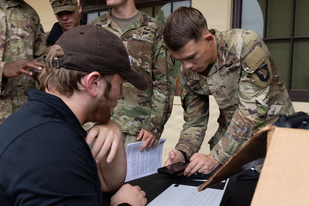Several U.S. Army soldiers in MultiCam/OCP camouflage uniforms are gathered around a table interacting with a civilian in a black cap and jacket. The central soldier, wearing a 101st Airborne Division patch and Airborne tab, is leaning over a laptop and smartphone on the table. The scene appears to involve administrative or check-in processing, with a document visible in the soldier's hand.