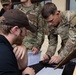 Several U.S. Army soldiers in MultiCam/OCP camouflage uniforms are gathered around a table interacting with a civilian in a black cap and jacket. The central soldier, wearing a 101st Airborne Division patch and Airborne tab, is leaning over a laptop and smartphone on the table. The scene appears to involve administrative or check-in processing, with a document visible in the soldier's hand.