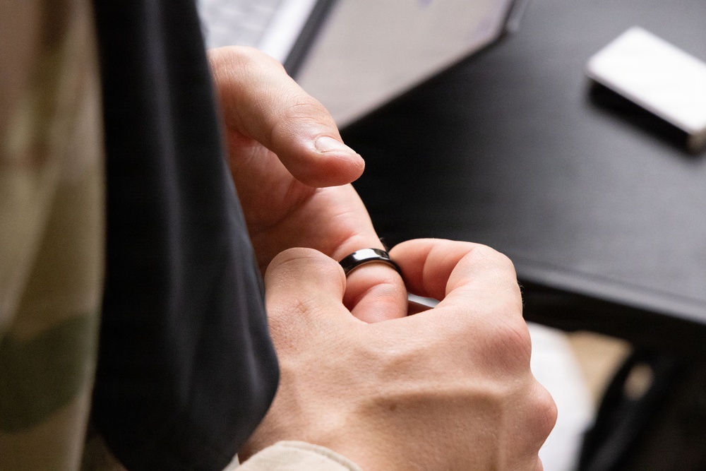 A person is handling a dark-colored ring, appearing to either place it on or remove it from a finger. The scene is set near a dark desk surface with a smartphone and what appears to be a laptop visible in the background. The focus is tightly on the hands and ring interaction.