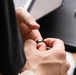 A person is handling a dark-colored ring, appearing to either place it on or remove it from a finger. The scene is set near a dark desk surface with a smartphone and what appears to be a laptop visible in the background. The focus is tightly on the hands and ring interaction.
