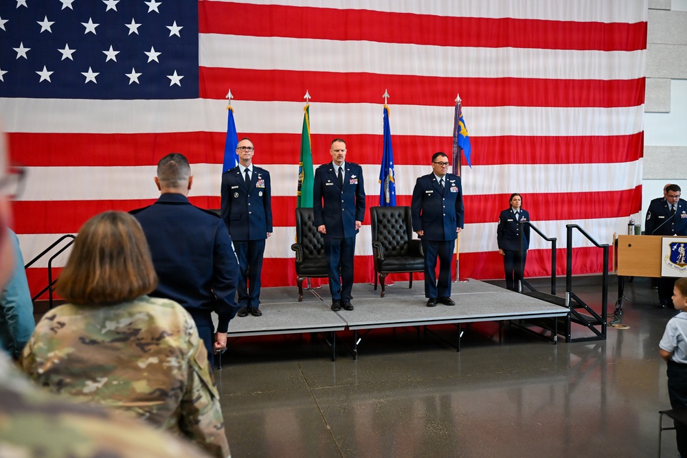 A formal U.S. Air Force military ceremony is taking place on a raised stage in front of a large American flag backdrop. Three officers in dress blue uniforms stand at attention on the stage, with two black leather chairs positioned behind them, suggesting a change of command or similar official ceremony. An audience including uniformed military personnel and civilians observes from the foreground.