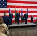 A formal U.S. Air Force military ceremony is taking place on a raised stage in front of a large American flag backdrop. Three officers in dress blue uniforms stand at attention on the stage, with two black leather chairs positioned behind them, suggesting a change of command or similar official ceremony. An audience including uniformed military personnel and civilians observes from the foreground.