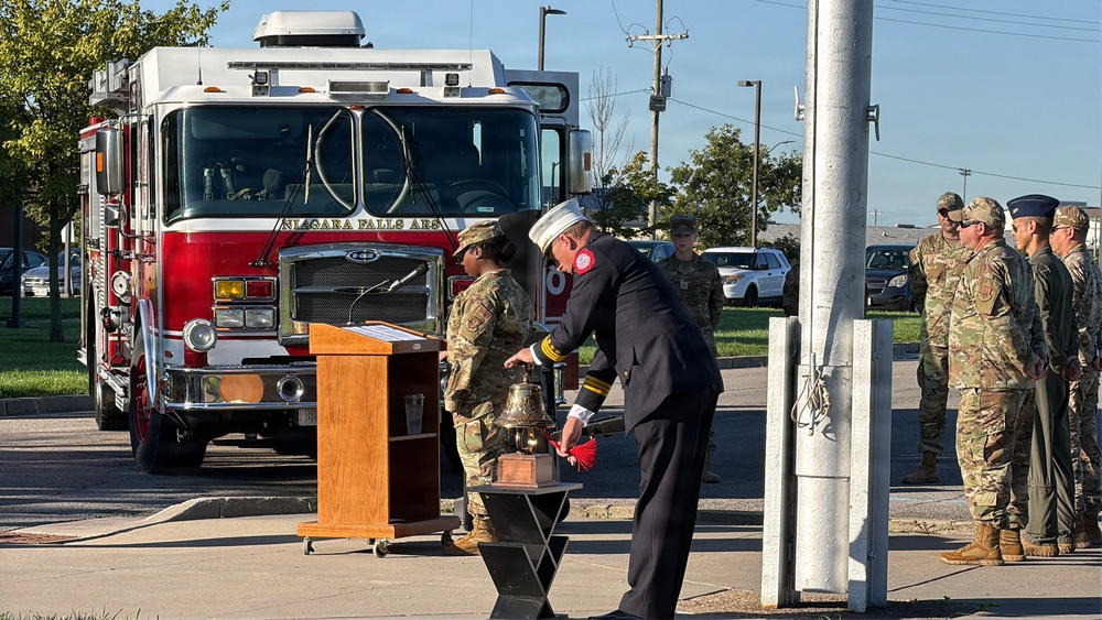 A formal outdoor ceremony is taking place at what appears to be Niagara Falls Air Reserve Station, featuring military personnel in camouflage uniforms and a fire official in dress uniform ringing a ceremonial bell. A red Niagara Falls ARS fire truck is prominently positioned in the background. The setting and bell-ringing ritual suggest this is likely a memorial or remembrance ceremony, possibly a 9/11 commemoration.