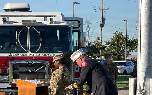 A formal outdoor ceremony is taking place at what appears to be Niagara Falls Air Reserve Station, featuring military personnel in camouflage uniforms and a fire official in dress uniform ringing a ceremonial bell. A red Niagara Falls ARS fire truck is prominently positioned in the background. The setting and bell-ringing ritual suggest this is likely a memorial or remembrance ceremony, possibly a 9/11 commemoration.