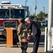 A formal outdoor ceremony is taking place at what appears to be Niagara Falls Air Reserve Station, featuring military personnel in camouflage uniforms and a fire official in dress uniform ringing a ceremonial bell. A red Niagara Falls ARS fire truck is prominently positioned in the background. The setting and bell-ringing ritual suggest this is likely a memorial or remembrance ceremony, possibly a 9/11 commemoration.