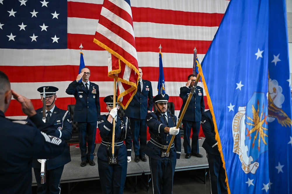 A formal United States Air Force color guard ceremony is taking place indoors, likely in a hangar or large ceremonial hall. Personnel in dress blue uniforms are presenting the American flag and the United States Air Force flag while officers on a raised stage render salutes. A large American flag serves as a backdrop, indicating a significant military ceremony such as a change of command or activation event.