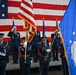 A formal United States Air Force color guard ceremony is taking place indoors, likely in a hangar or large ceremonial hall. Personnel in dress blue uniforms are presenting the American flag and the United States Air Force flag while officers on a raised stage render salutes. A large American flag serves as a backdrop, indicating a significant military ceremony such as a change of command or activation event.