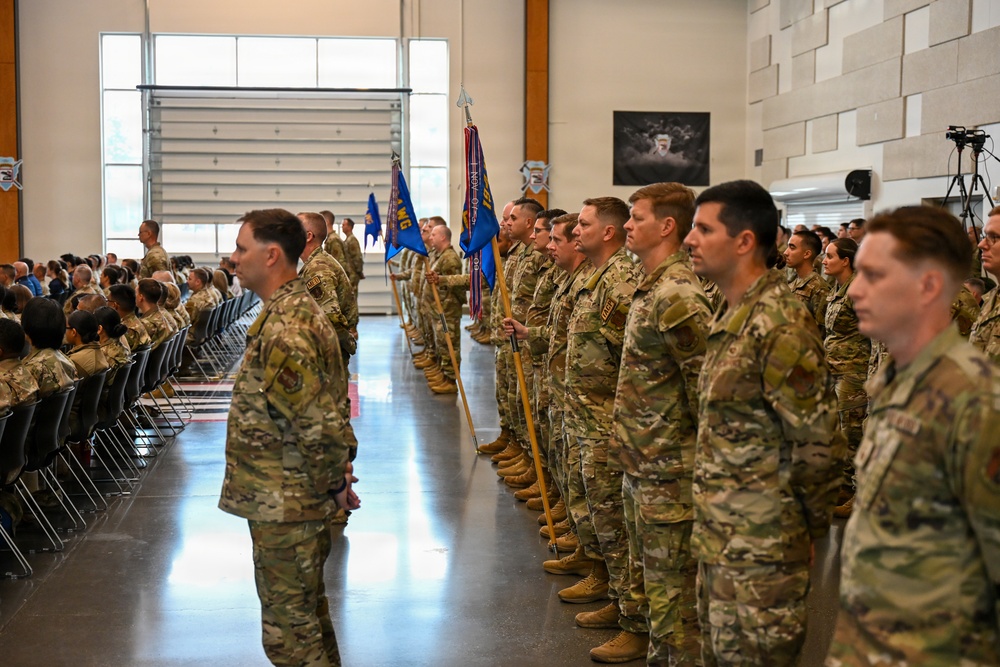 A formal military ceremony is taking place inside a large indoor facility, likely a hangar or armory. Personnel in MultiCam/OCP camouflage uniforms are standing in formation, with color guard members holding blue unit guidons/flags. A seated audience of additional military personnel and civilians observes from the left side.