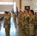 A formal military ceremony is taking place inside a large indoor facility, likely a hangar or armory. Personnel in MultiCam/OCP camouflage uniforms are standing in formation, with color guard members holding blue unit guidons/flags. A seated audience of additional military personnel and civilians observes from the left side.