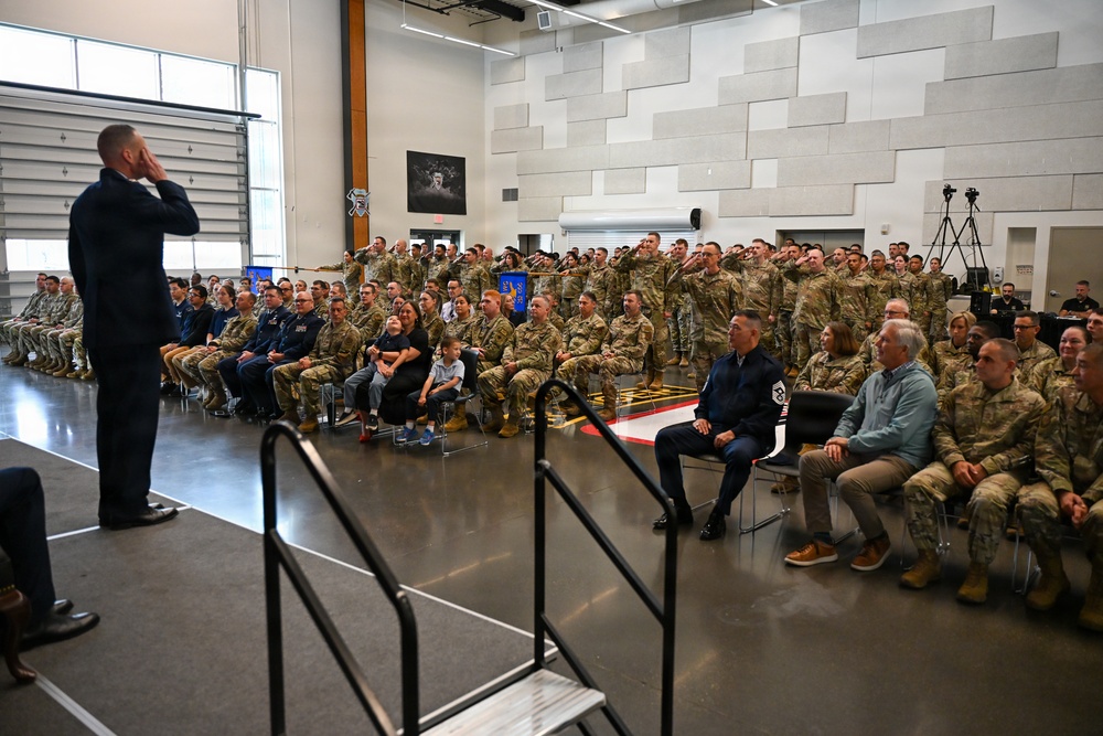 A military ceremony or formation is taking place inside a large indoor facility, likely a hangar or training bay. A uniformed officer on a raised stage is rendering a salute to a large assembled group of military personnel, many of whom are returning the salute. The audience includes a mix of uniformed service members in camouflage and dress uniforms, as well as a few civilians including children.