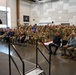 A military ceremony or formation is taking place inside a large indoor facility, likely a hangar or training bay. A uniformed officer on a raised stage is rendering a salute to a large assembled group of military personnel, many of whom are returning the salute. The audience includes a mix of uniformed service members in camouflage and dress uniforms, as well as a few civilians including children.