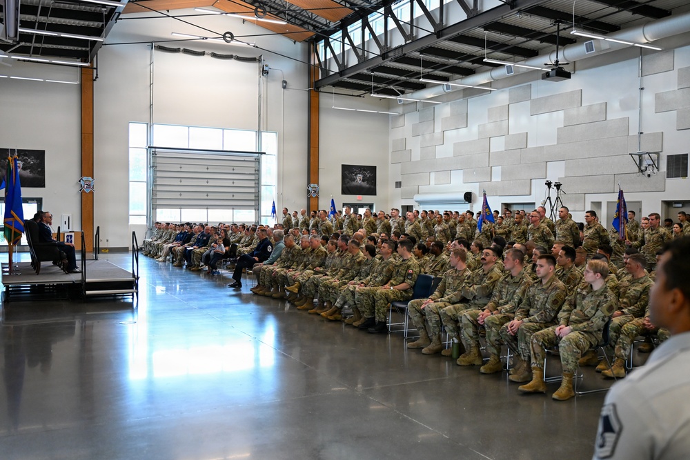 A large military formation is seated in rows inside a modern hangar or multipurpose facility, facing a raised stage where officials are seated. The gathering appears to be a formal military ceremony or briefing, with personnel dressed in camouflage uniforms and a small number in dress blues. Military flags/guidons are visible throughout the space.