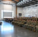 A large military formation is seated in rows inside a modern hangar or multipurpose facility, facing a raised stage where officials are seated. The gathering appears to be a formal military ceremony or briefing, with personnel dressed in camouflage uniforms and a small number in dress blues. Military flags/guidons are visible throughout the space.