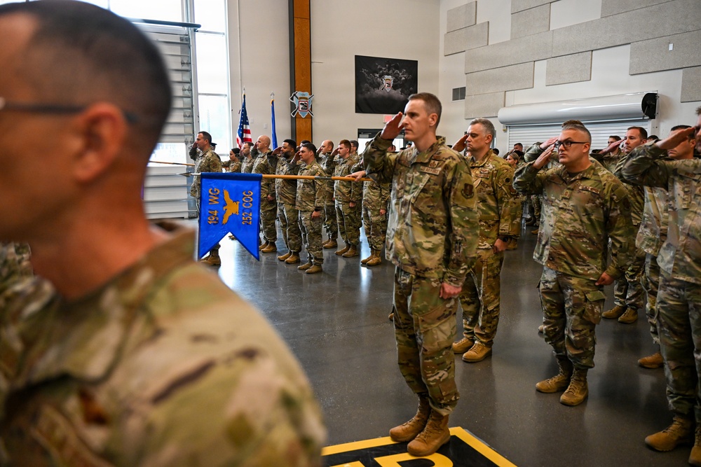 Military personnel in OCP (Operational Camouflage Pattern) uniforms are standing at attention and rendering salutes during a formal ceremony indoors. A blue guidon pennant is visible bearing the markings '194 WG' and '252 COG', indicating this is an Air National Guard unit formation. The ceremony appears to be taking place inside a hangar or large military facility.