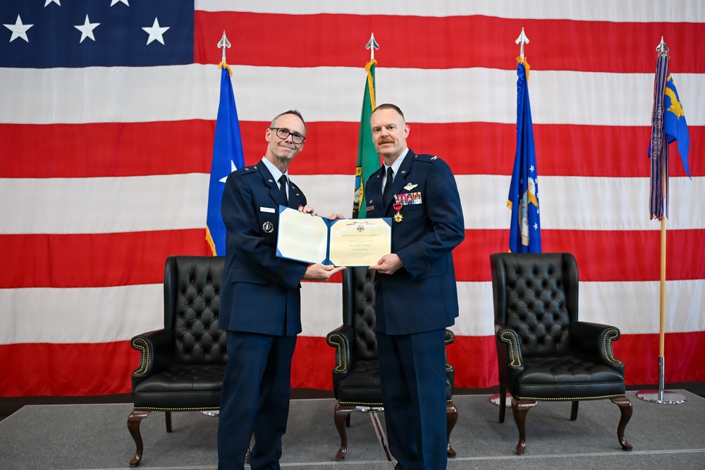 Two U.S. Air Force officers in dress blue uniforms are posing together while jointly holding an official certificate or award document during a formal military ceremony. The setting features a large American flag as a backdrop along with several military and organizational flags. The officer on the right displays multiple service ribbons and medals, suggesting a retirement, promotion, or commendation ceremony.