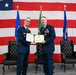 Two U.S. Air Force officers in dress blue uniforms are posing together while jointly holding an official certificate or award document during a formal military ceremony. The setting features a large American flag as a backdrop along with several military and organizational flags. The officer on the right displays multiple service ribbons and medals, suggesting a retirement, promotion, or commendation ceremony.