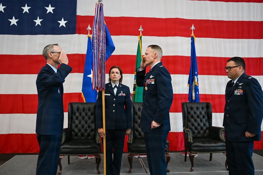 A formal U.S. Air Force change of command or promotion ceremony is taking place in front of a large American flag. Two officers are exchanging salutes while a female airman holds a unit guidon/flag with streamers, and a fourth officer stands at attention to the right. The setting includes leather chairs on a stage, suggesting an official military ceremony.
