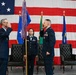 A formal U.S. Air Force change of command or promotion ceremony is taking place in front of a large American flag. Two officers are exchanging salutes while a female airman holds a unit guidon/flag with streamers, and a fourth officer stands at attention to the right. The setting includes leather chairs on a stage, suggesting an official military ceremony.