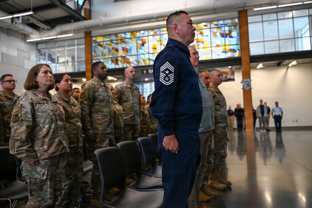 A military formation or ceremony is taking place inside a modern indoor facility, likely a military installation or base. A senior non-commissioned officer in a blue Air Force uniform with a Command Chief Master Sergeant insignia stands prominently in the foreground, while numerous personnel in OCP (Operational Camouflage Pattern) uniforms stand in formation behind him. The setting features a distinctive stained glass window backdrop and modern architecture.