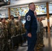 A military formation or ceremony is taking place inside a modern indoor facility, likely a military installation or base. A senior non-commissioned officer in a blue Air Force uniform with a Command Chief Master Sergeant insignia stands prominently in the foreground, while numerous personnel in OCP (Operational Camouflage Pattern) uniforms stand in formation behind him. The setting features a distinctive stained glass window backdrop and modern architecture.