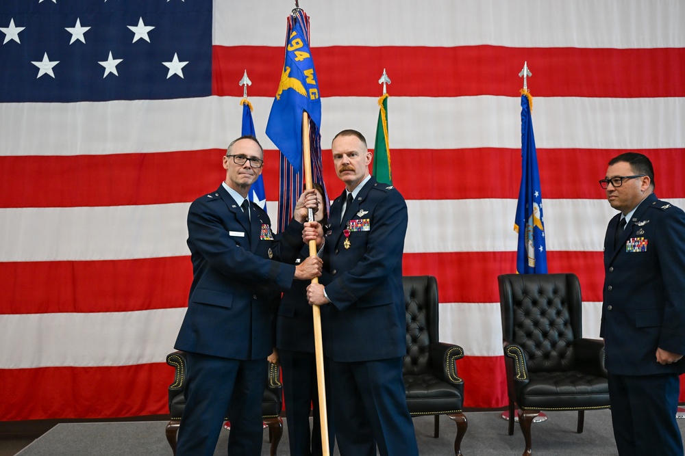 A military change of command ceremony is taking place in front of a large American flag backdrop. Two U.S. Air Force officers in dress blue uniforms are jointly holding a unit guidon (flag on a staff), which is the central symbolic act of a change of command. A third officer stands at attention in the background to the right.