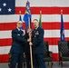 A military change of command ceremony is taking place in front of a large American flag backdrop. Two U.S. Air Force officers in dress blue uniforms are jointly holding a unit guidon (flag on a staff), which is the central symbolic act of a change of command. A third officer stands at attention in the background to the right.