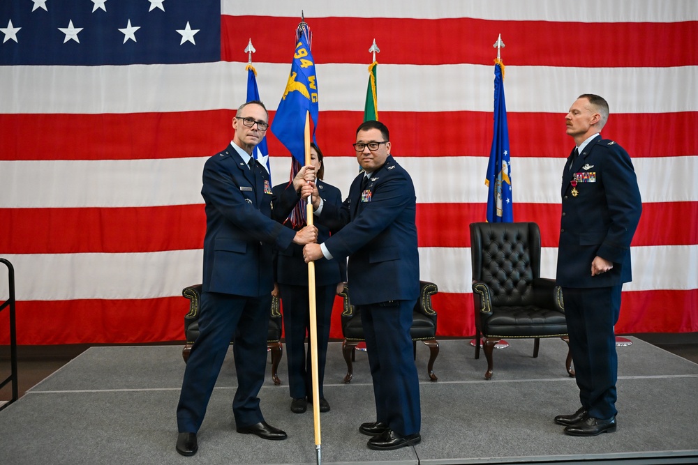 A formal U.S. Air Force change of command ceremony is taking place on a stage in front of a large American flag backdrop. Two officers in dress blue uniforms are exchanging a unit guidon (flag on a staff), which is the traditional centerpiece of a change of command ceremony. Additional personnel including a third officer and a flag bearer are visible on stage.