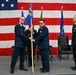 A formal U.S. Air Force change of command ceremony is taking place on a stage in front of a large American flag backdrop. Two officers in dress blue uniforms are exchanging a unit guidon (flag on a staff), which is the traditional centerpiece of a change of command ceremony. Additional personnel including a third officer and a flag bearer are visible on stage.