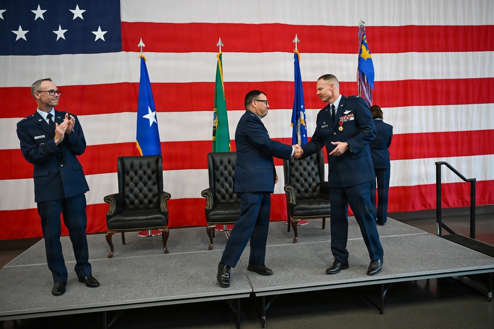A formal U.S. Air Force military ceremony is taking place on a stage in front of a large American flag backdrop. Two officers in dress blue uniforms are shaking hands at center stage while a third officer applauds from the left. The presence of formal chairs, multiple flags, and military decorations suggests this is a change of command or award ceremony.