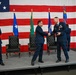 A formal U.S. Air Force military ceremony is taking place on a stage in front of a large American flag backdrop. Two officers in dress blue uniforms are shaking hands at center stage while a third officer applauds from the left. The presence of formal chairs, multiple flags, and military decorations suggests this is a change of command or award ceremony.