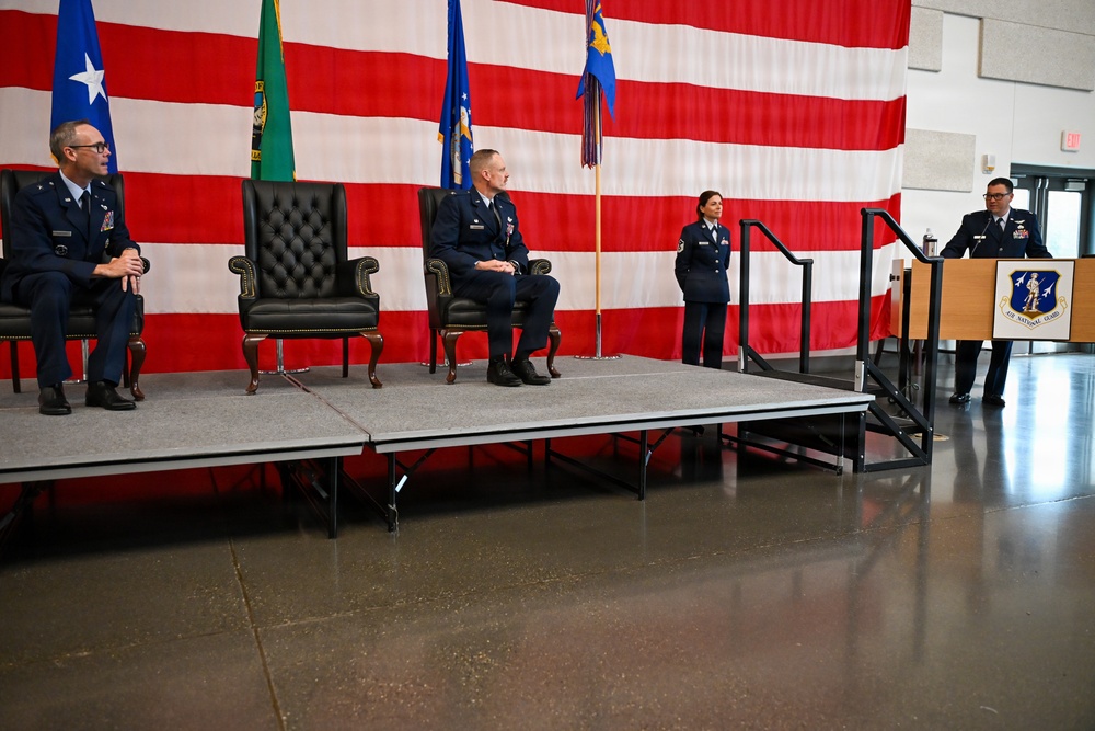 A formal Air National Guard military ceremony is taking place in what appears to be a hangar or large indoor facility. Two officers are seated in ceremonial chairs on a raised stage platform while a third officer speaks at a podium bearing the Air National Guard insignia. An American flag backdrop, Washington State flag, and military unit flags are prominently displayed.