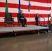 A formal Air National Guard military ceremony is taking place in what appears to be a hangar or large indoor facility. Two officers are seated in ceremonial chairs on a raised stage platform while a third officer speaks at a podium bearing the Air National Guard insignia. An American flag backdrop, Washington State flag, and military unit flags are prominently displayed.