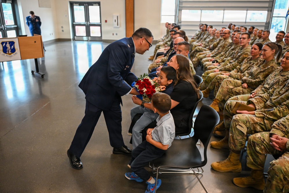 A military ceremony is taking place in what appears to be an Air National Guard facility. An officer in dress blue uniform is presenting a bouquet of red flowers to a woman seated with two young children, likely a family recognition moment during a formal military event. Rows of uniformed personnel in camouflage (OCP) uniforms are seated as an audience.