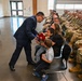 A military ceremony is taking place in what appears to be an Air National Guard facility. An officer in dress blue uniform is presenting a bouquet of red flowers to a woman seated with two young children, likely a family recognition moment during a formal military event. Rows of uniformed personnel in camouflage (OCP) uniforms are seated as an audience.