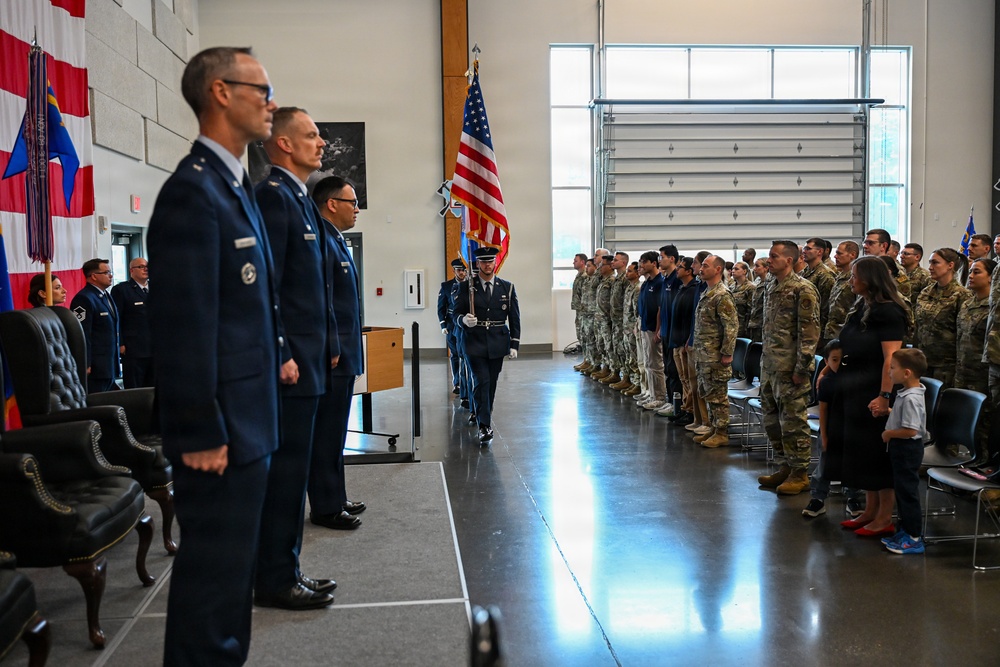 A formal military ceremony is taking place inside a large indoor facility, likely a hangar or event hall. A color guard member is marching with the American flag while officers in dress blues and troops in combat uniforms stand at attention on either side. The scene appears to be a change of command, activation, or similar official military ceremony.