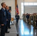 A formal military ceremony is taking place inside a large indoor facility, likely a hangar or event hall. A color guard member is marching with the American flag while officers in dress blues and troops in combat uniforms stand at attention on either side. The scene appears to be a change of command, activation, or similar official military ceremony.