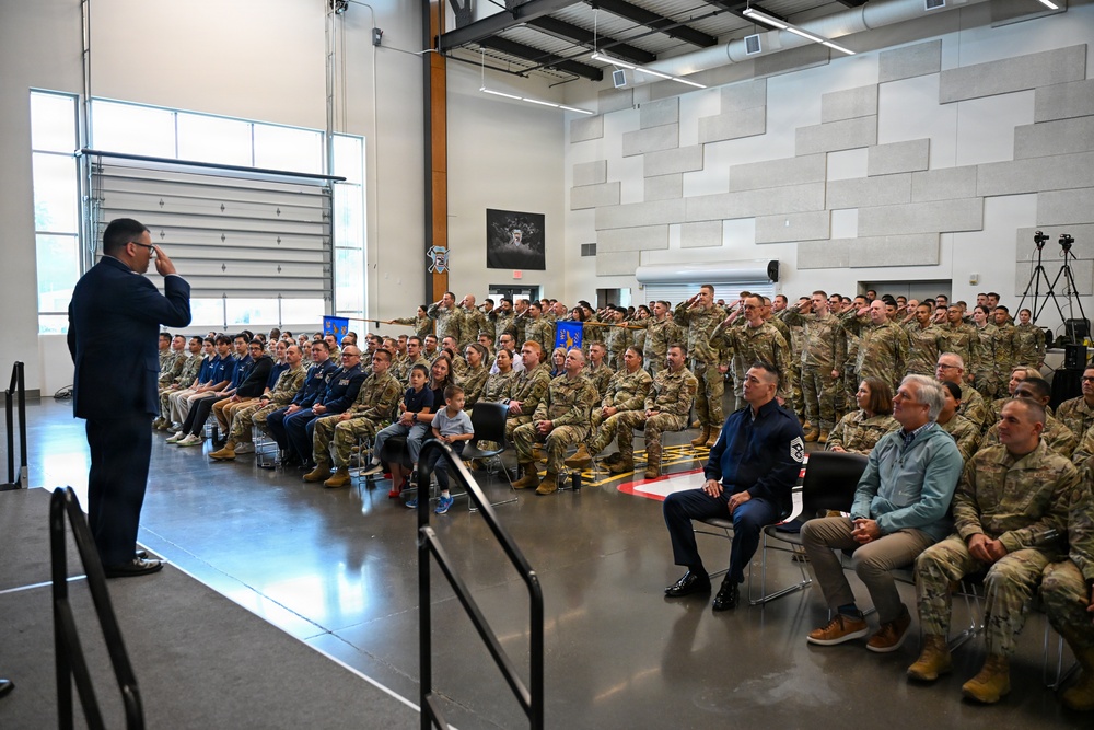 A military ceremony or formation is taking place inside a large hangar-style facility. A uniformed officer stands on a raised platform rendering a salute to an assembled audience of military personnel and civilians. Many attendees in the back rows are returning the salute while front rows remain seated.