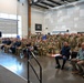 A military ceremony or formation is taking place inside a large hangar-style facility. A uniformed officer stands on a raised platform rendering a salute to an assembled audience of military personnel and civilians. Many attendees in the back rows are returning the salute while front rows remain seated.