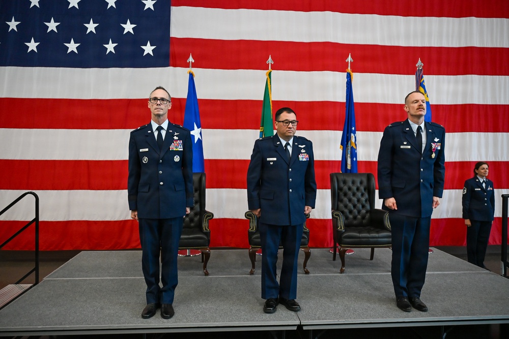 Three United States Air Force officers stand at attention on a stage during a formal military ceremony, likely a change of command. A large American flag serves as the backdrop, flanked by multiple military unit flags on staffs. Two ceremonial chairs are positioned behind the officers on the stage.