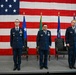 Three United States Air Force officers stand at attention on a stage during a formal military ceremony, likely a change of command. A large American flag serves as the backdrop, flanked by multiple military unit flags on staffs. Two ceremonial chairs are positioned behind the officers on the stage.
