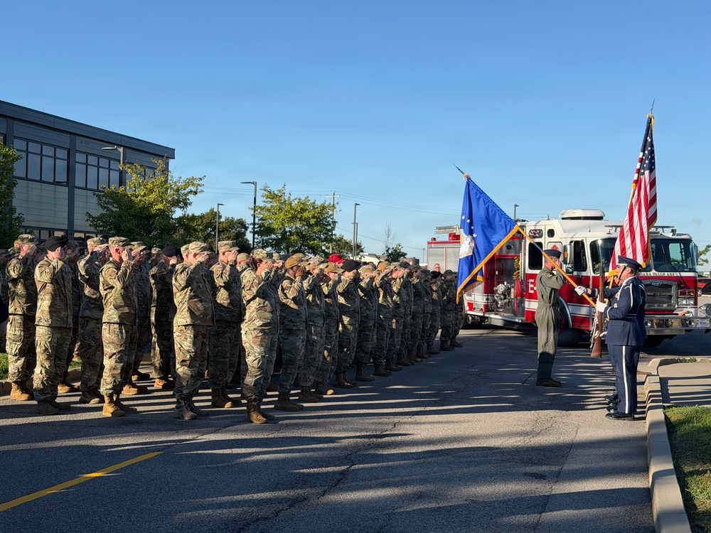 A large formation of military personnel in camouflage uniforms are rendering a salute during a formal outdoor ceremony. A color guard is presenting the United States flag and what appears to be an Air Force unit flag, with a fire truck visible in the background. The ceremony appears to be taking place in a parking area near a modern building, likely on a military installation.