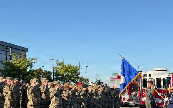 A large formation of military personnel in camouflage uniforms are rendering a salute during a formal outdoor ceremony. A color guard is presenting the United States flag and what appears to be an Air Force unit flag, with a fire truck visible in the background. The ceremony appears to be taking place in a parking area near a modern building, likely on a military installation.