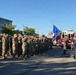 A large formation of military personnel in camouflage uniforms are rendering a salute during a formal outdoor ceremony. A color guard is presenting the United States flag and what appears to be an Air Force unit flag, with a fire truck visible in the background. The ceremony appears to be taking place in a parking area near a modern building, likely on a military installation.