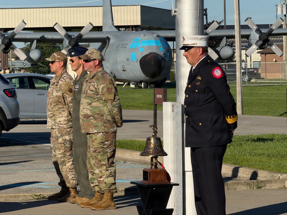 A solemn outdoor ceremony is taking place at what appears to be a military air base, featuring uniformed Air Force personnel in camouflage uniforms alongside a fire department official in dress uniform. A ceremonial brass bell on a wooden base is positioned near a white pole, suggesting a memorial or remembrance ceremony. A static display C-130 Hercules aircraft with 'AIR FORCE' markings is visible in the background.