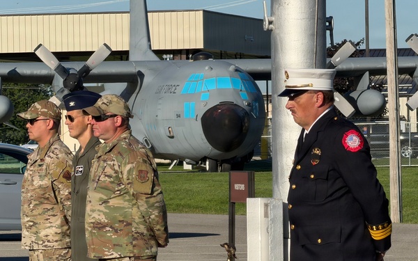 A solemn outdoor ceremony is taking place at what appears to be a military air base, featuring uniformed Air Force personnel in camouflage uniforms alongside a fire department official in dress uniform. A ceremonial brass bell on a wooden base is positioned near a white pole, suggesting a memorial or remembrance ceremony. A static display C-130 Hercules aircraft with 'AIR FORCE' markings is visible in the background.