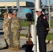 A solemn outdoor ceremony is taking place at what appears to be a military air base, featuring uniformed Air Force personnel in camouflage uniforms alongside a fire department official in dress uniform. A ceremonial brass bell on a wooden base is positioned near a white pole, suggesting a memorial or remembrance ceremony. A static display C-130 Hercules aircraft with 'AIR FORCE' markings is visible in the background.