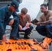 Three military personnel are gathered on the deck of a naval vessel conducting what appears to be a tool inventory or inspection. An orange tool roll is spread out on the deck displaying various hand tools, while one person references a document and another takes notes. The setting is clearly a ship's exterior deck with naval equipment visible in the background.