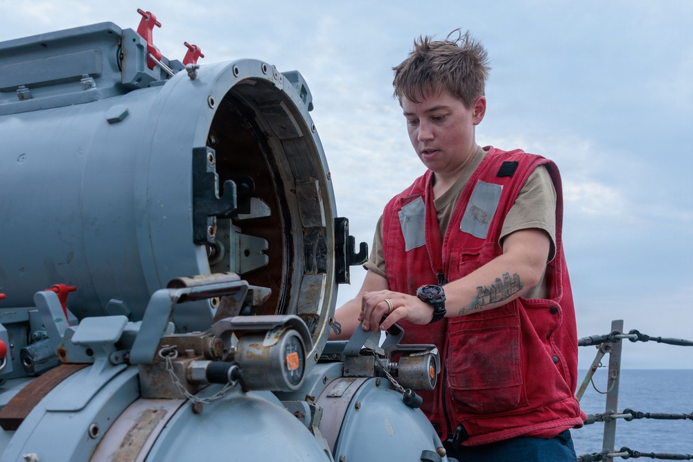 A person wearing a red vest and tan shirt is working on what appears to be a naval weapon system or torpedo tube aboard a military vessel at sea. The individual has short brown hair, visible tattoos on their forearm, and is wearing a watch while manipulating components of the equipment. The ocean and overcast sky are visible in the background.
